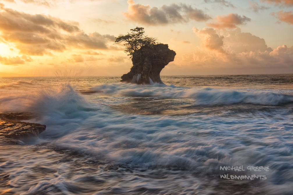 The stoic profile of a sea stack contrasts with the fading light of dusk amid a turbulent Caribbean surf and colorful evening...