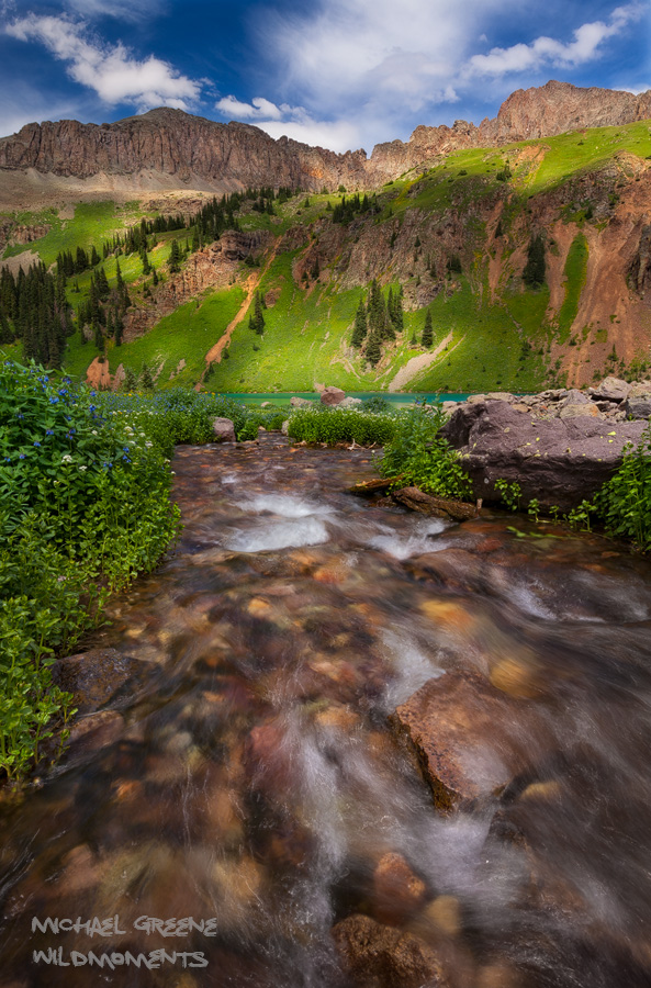 Lower Blue Lake during peak wildflower season as seen from a tributary on a partly cloudy day. Find this lake near the town of...