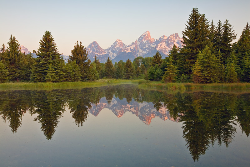 The soft light of sunrise illuminates the Teton peaks still covered in snow above 8500 feet reflected in the launching waters...